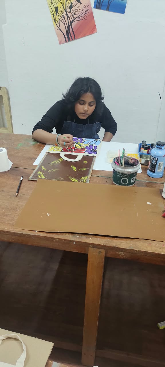 A student carefully painting a floral design onto a canvas tote bag at the art table