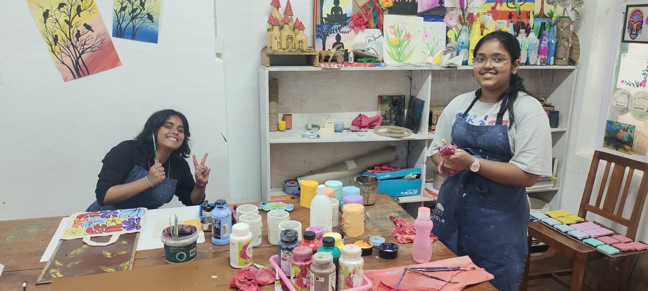 Two Art Elective students smiling with their painted tote bag and the studio's paint supplies